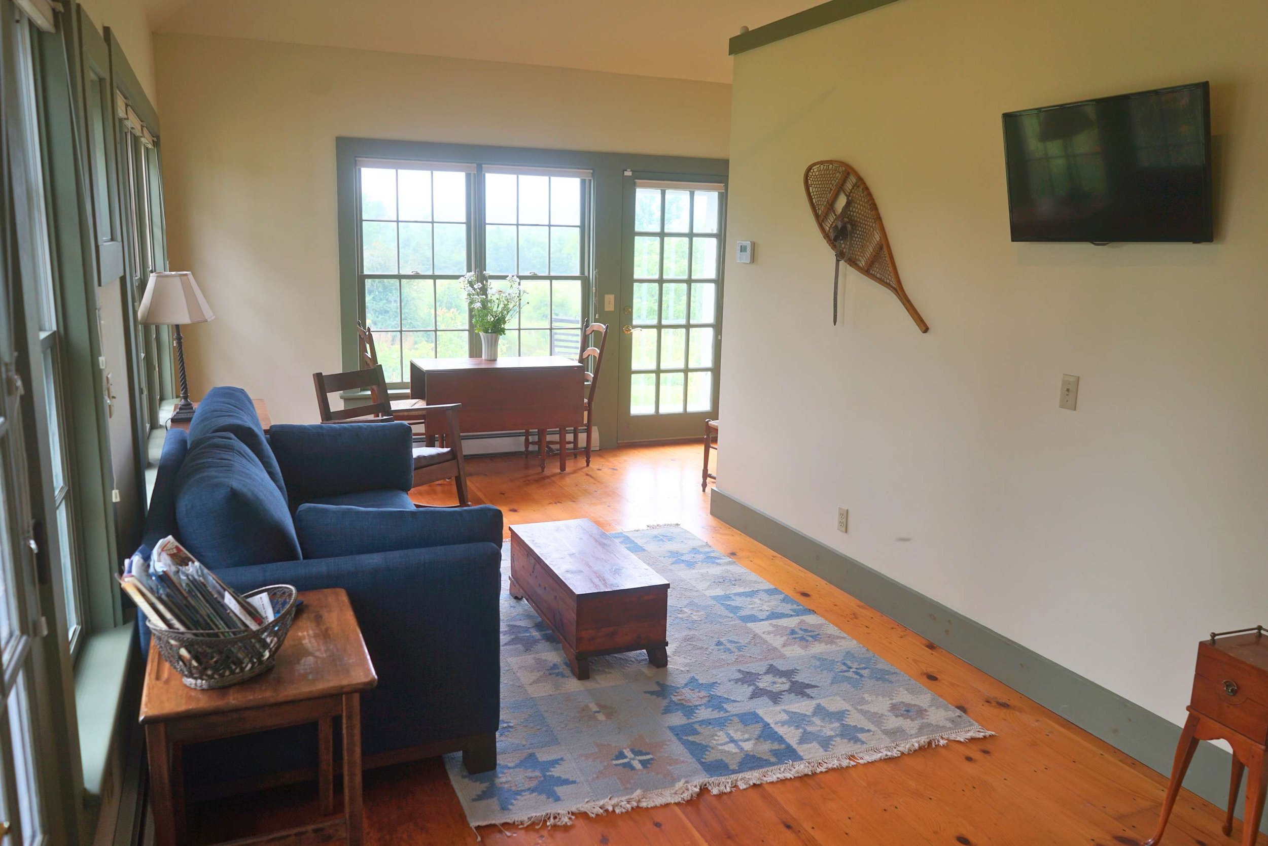 Living room with blue sofa, wooden coffee table, and a small side table with a basket of magazines. There is a wood dining table with chairs by large windows and a glass door leading outside. A mounted snowshoe and a flat-screen TV are on the wall.